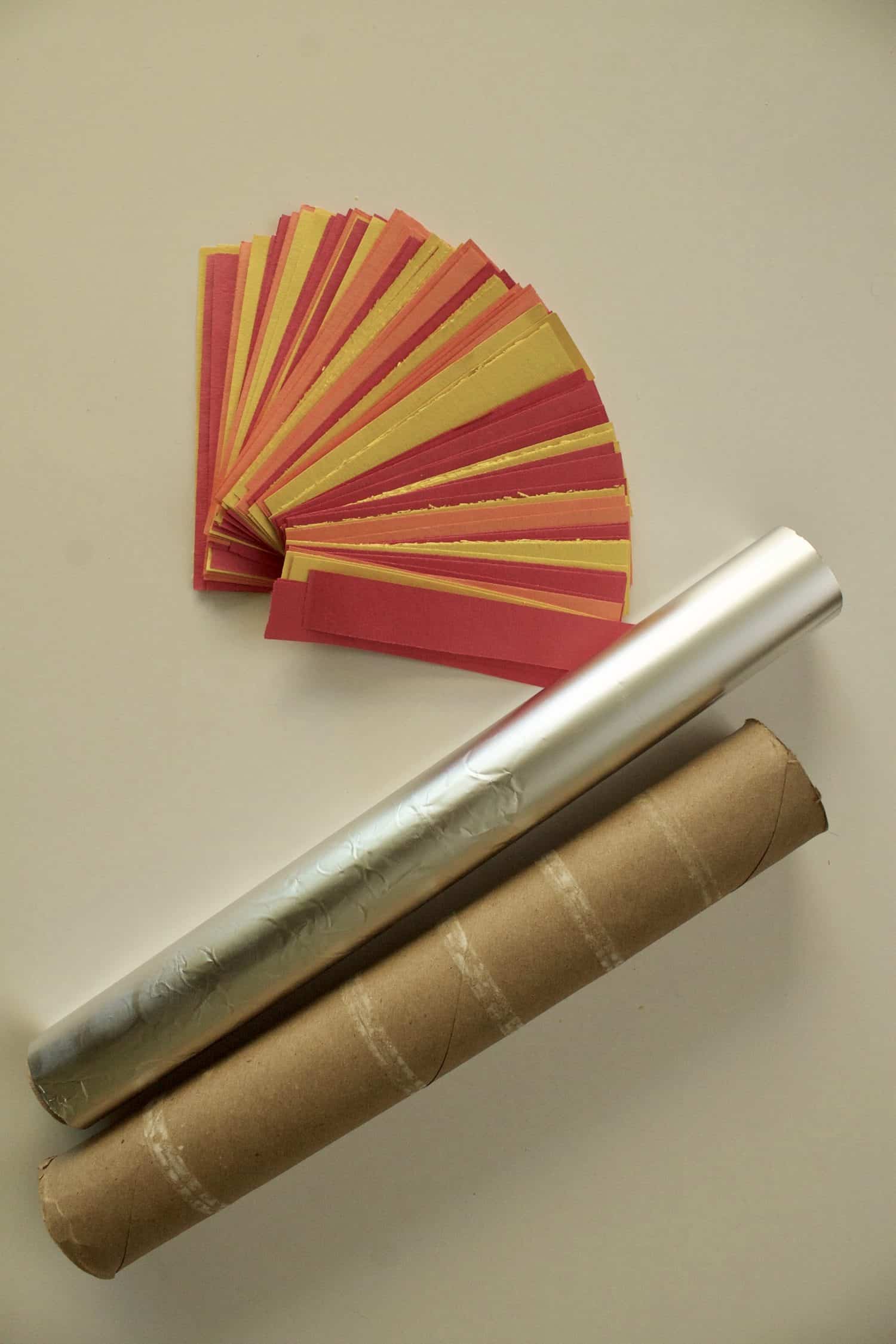 a stack of small strips of yellow, red, and orange construction paper, a roll of aluminum foil, and a cardboard tube